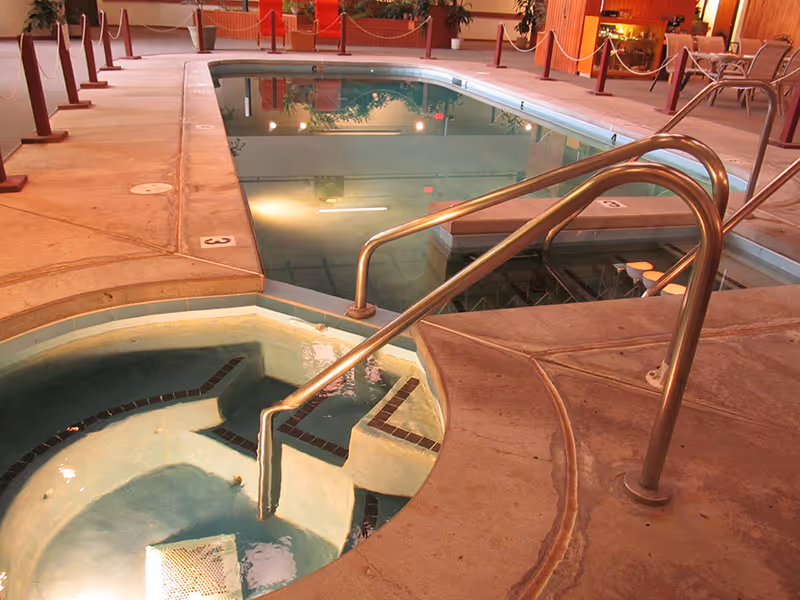 Indoor pool area with a hot tub in the foreground and a swimming pool in the background. The area is surrounded by a concrete deck with metal handrails and some seating visible in the distance.