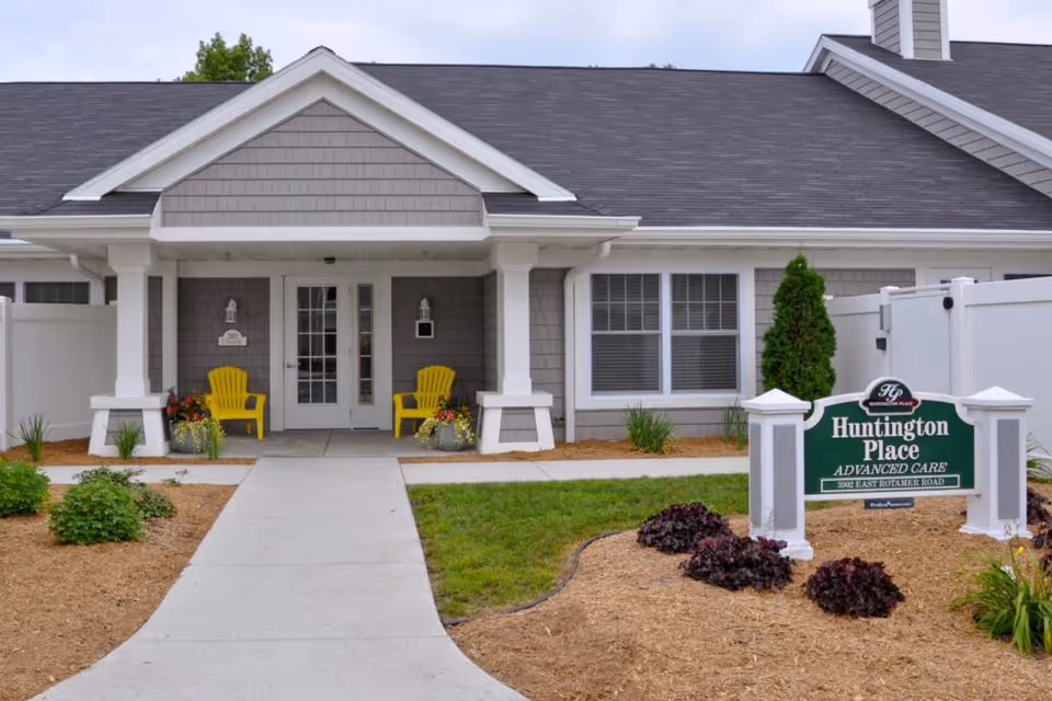 Front exterior view of Huntington Place Advanced Care facility with a concrete walkway leading to a covered entrance. Two yellow chairs are placed on either side of the door, surrounded by potted plants. The building has gray siding and white trim, with a green sign in the landscaped front yard displaying the facility name and address.