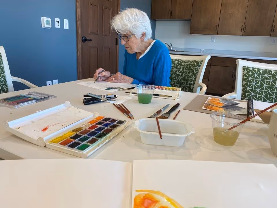 An elderly woman with white hair and glasses is sitting at a table in a room with wooden cabinets and a blue wall. She is painting with watercolors, surrounded by art supplies including paintbrushes, watercolor palettes, and cups of water. The table has several sheets of paper with paintings in progress.