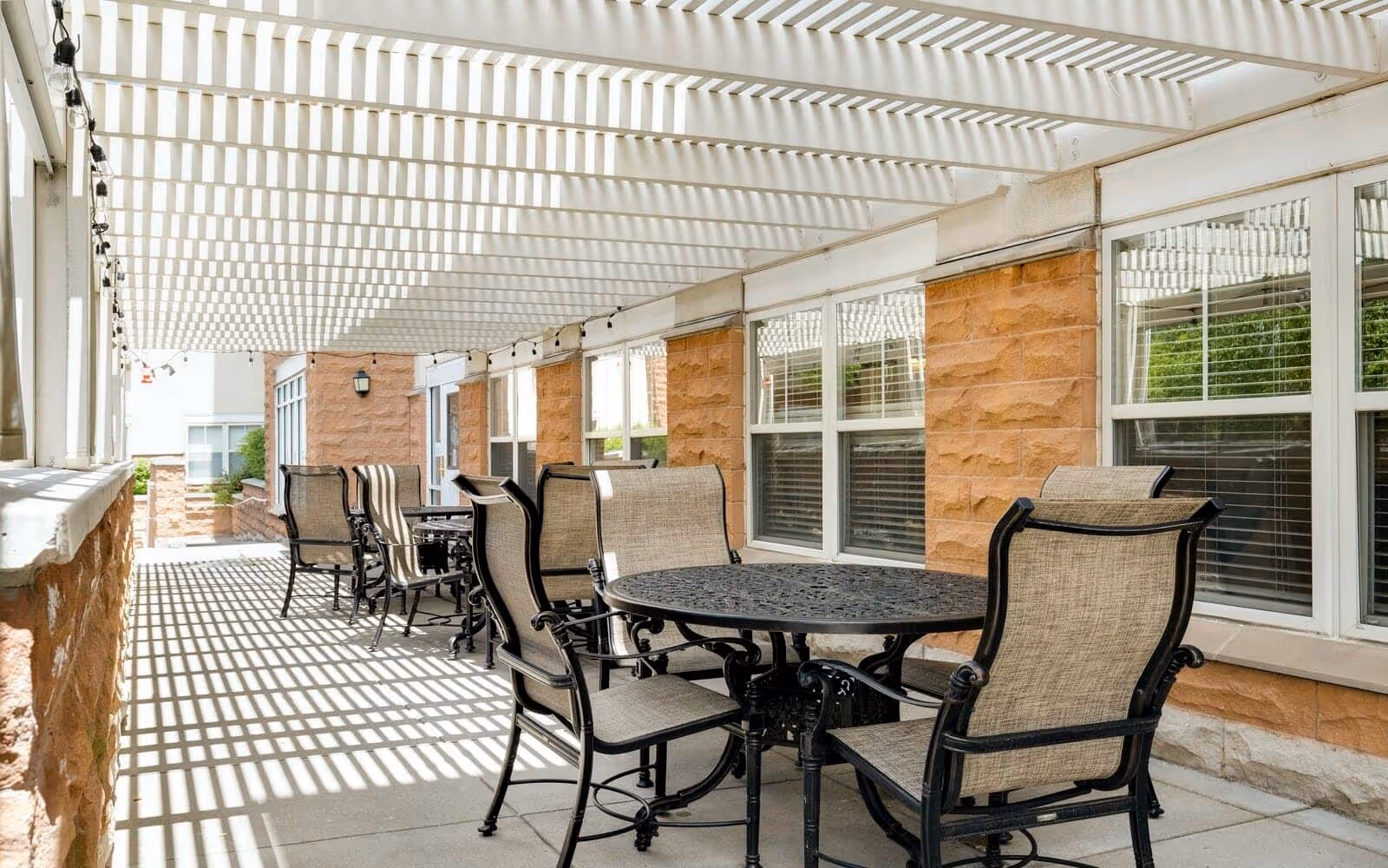 Outdoor covered patio area with multiple black metal tables and beige cushioned chairs. The patio has a white pergola overhead casting striped shadows on the floor and furniture. The building wall is made of reddish-brown stone with white-framed windows.