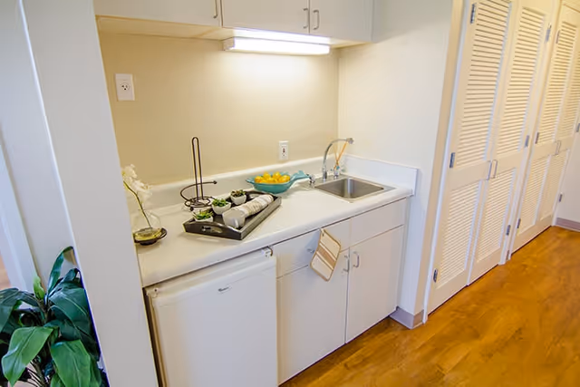 Small kitchenette with white cabinets, a sink, mini refrigerator and countertop accessories in a residential unit.