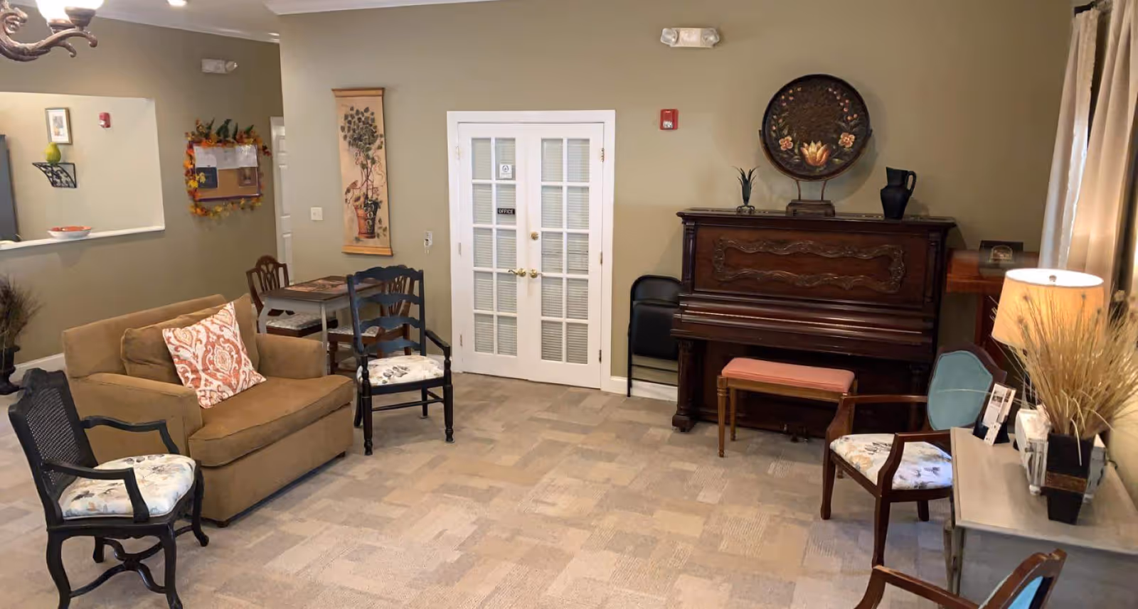Communal living room with a sofa and several chairs arranged around an upright piano and French doors.