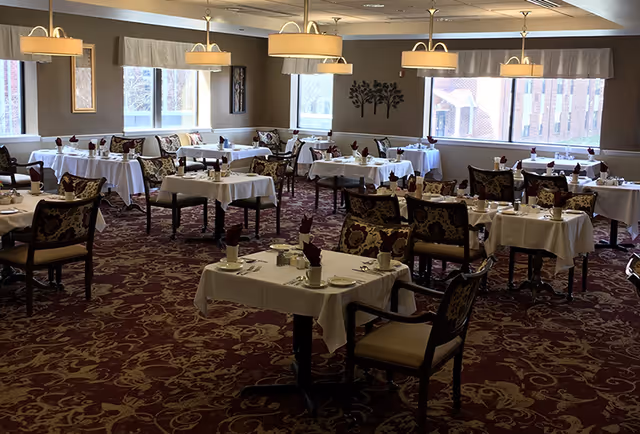A dining room with multiple tables covered in white tablecloths, each set with cups, plates, napkins, and silverware. The room has patterned carpet, upholstered chairs with floral designs, large windows letting in natural light, and ceiling lights with beige lampshades.