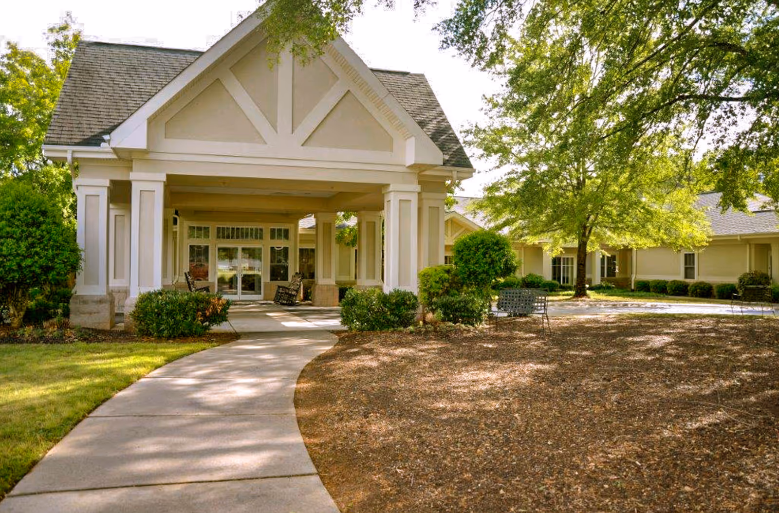 Entrance of The Garden House of Anderson facility with a covered porch supported by columns, a curved concrete walkway leading to double glass doors, surrounded by green bushes, trees, and outdoor seating.