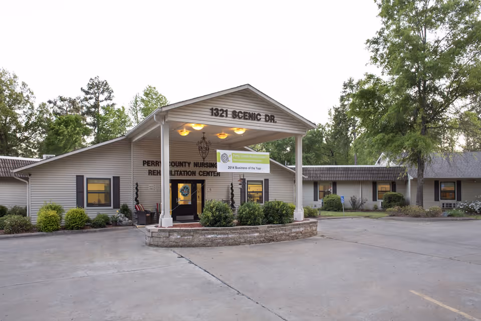 Front exterior of Perry County Nursing & Rehabilitation Center showing the covered entrance with address '1321 Scenic Dr', landscaped beds, and parking area.