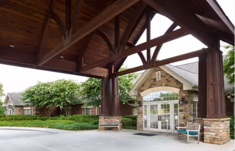 Covered entrance area of a senior living facility with a large wooden canopy supported by thick wooden beams. There are two benches on either side of the entrance door. The building exterior features stone and brickwork with large glass double doors. Green shrubs and trees surround the entrance.