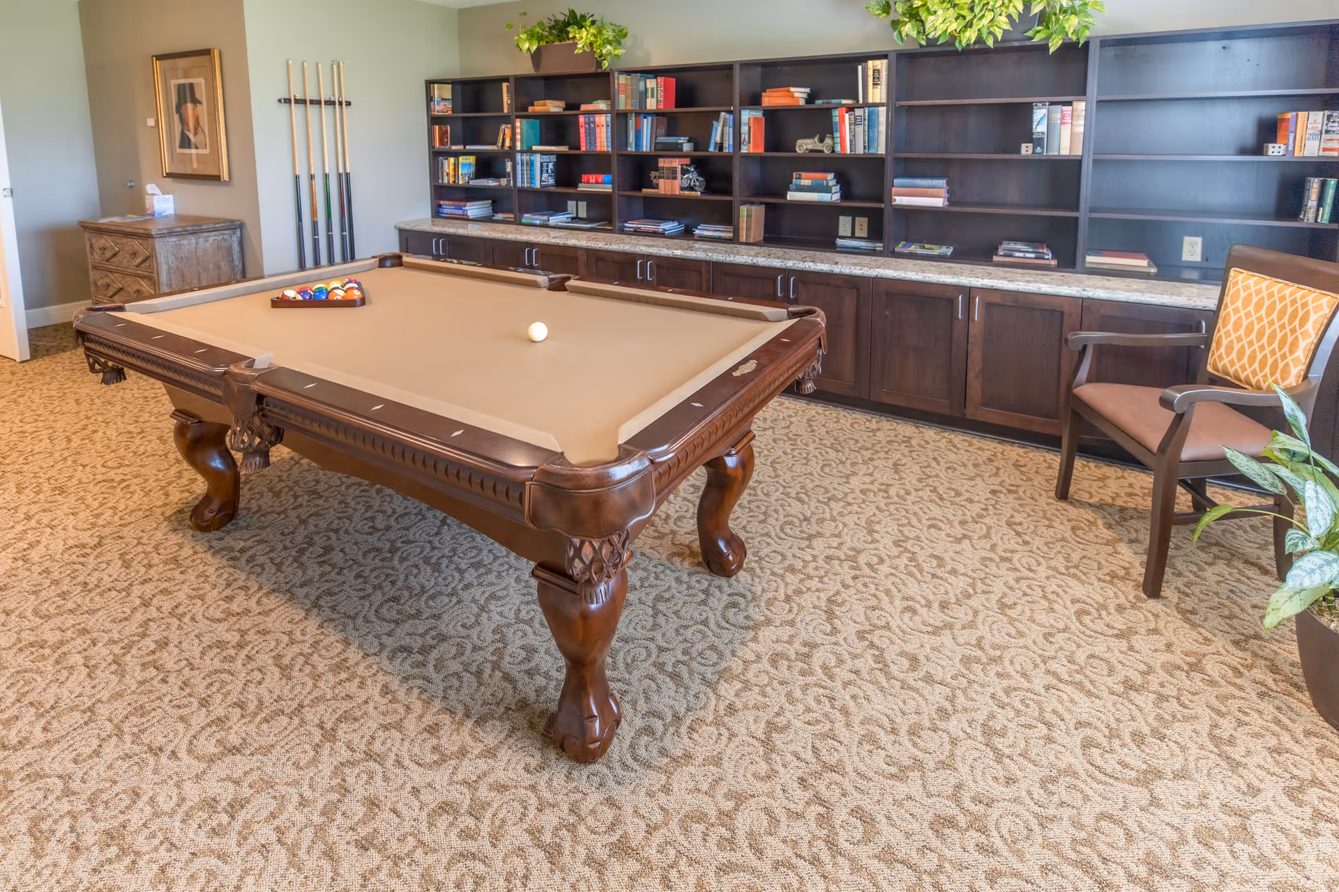 A room with a beige carpet featuring a patterned design, a brown pool table with billiard balls arranged on it, a wooden chair with an orange patterned cushion, a large dark wood bookshelf filled with books and decorative items, and a small chest of drawers with a framed picture above it. There are also pool cues standing against the wall and some green plants on top of the bookshelf.