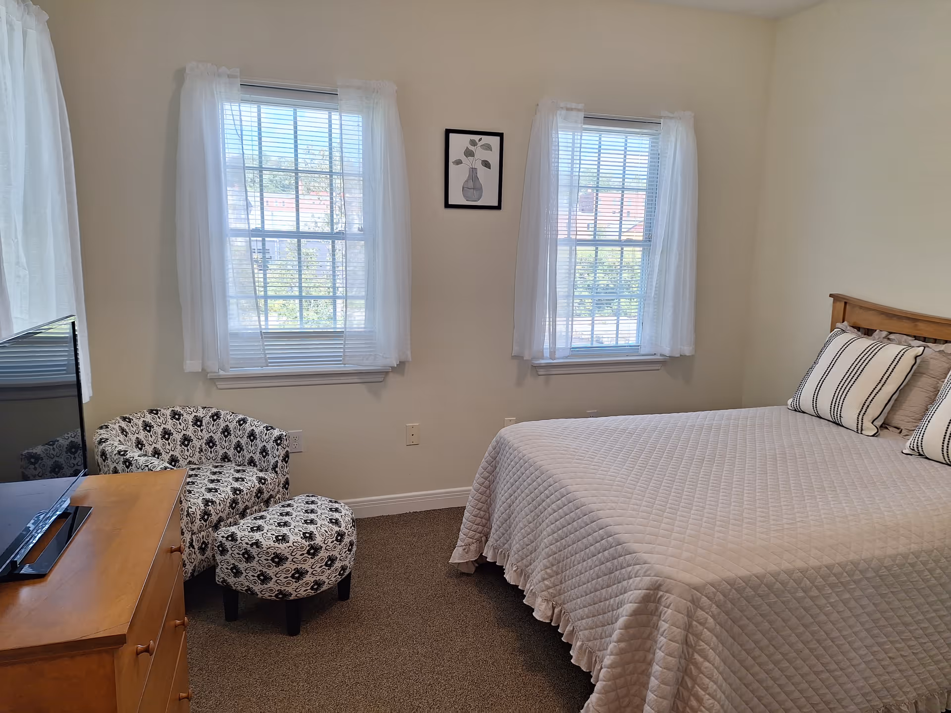 A bedroom with a bed covered in a light beige quilt and two striped pillows. There are two windows with white sheer curtains on the wall behind the bed. A wooden dresser with a flat-screen TV on top is positioned to the left, next to a patterned armchair and matching ottoman. A framed picture of a vase with leaves hangs between the windows.