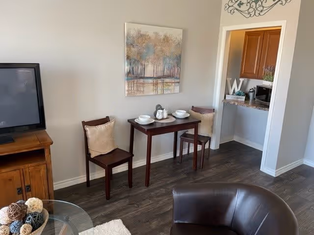 A cozy living area with a small wooden table set for two with white dishes and two wooden chairs with beige cushions. A flat-screen TV sits on a wooden cabinet to the left. A glass coffee table with decorative balls is partially visible in the foreground. The floor is dark wood, and a painting of trees hangs on the wall above the table. To the right, there is an open doorway leading to a small kitchen area with wooden cabinets and a microwave.