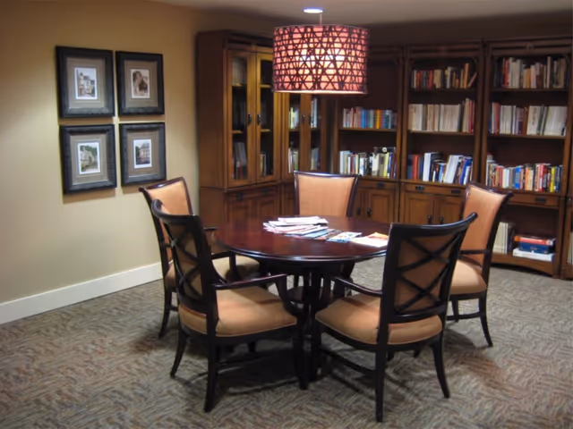 A cozy reading or meeting area with a round wooden table surrounded by five cushioned chairs. Behind the table are wooden bookshelves filled with books and a cabinet with glass doors. A decorative hanging lamp with a patterned shade is above the table, and four framed pictures hang on the wall to the left.