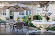 Stone tiered fountain in a sunny courtyard with patio tables, chairs, an umbrella, and a building in the background.