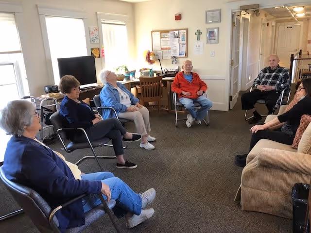 A group of elderly people sitting in a circle in a well-lit room with windows, chairs, a television, a desk with a lamp, and a bulletin board on the wall. The room has carpeted flooring and light-colored walls.