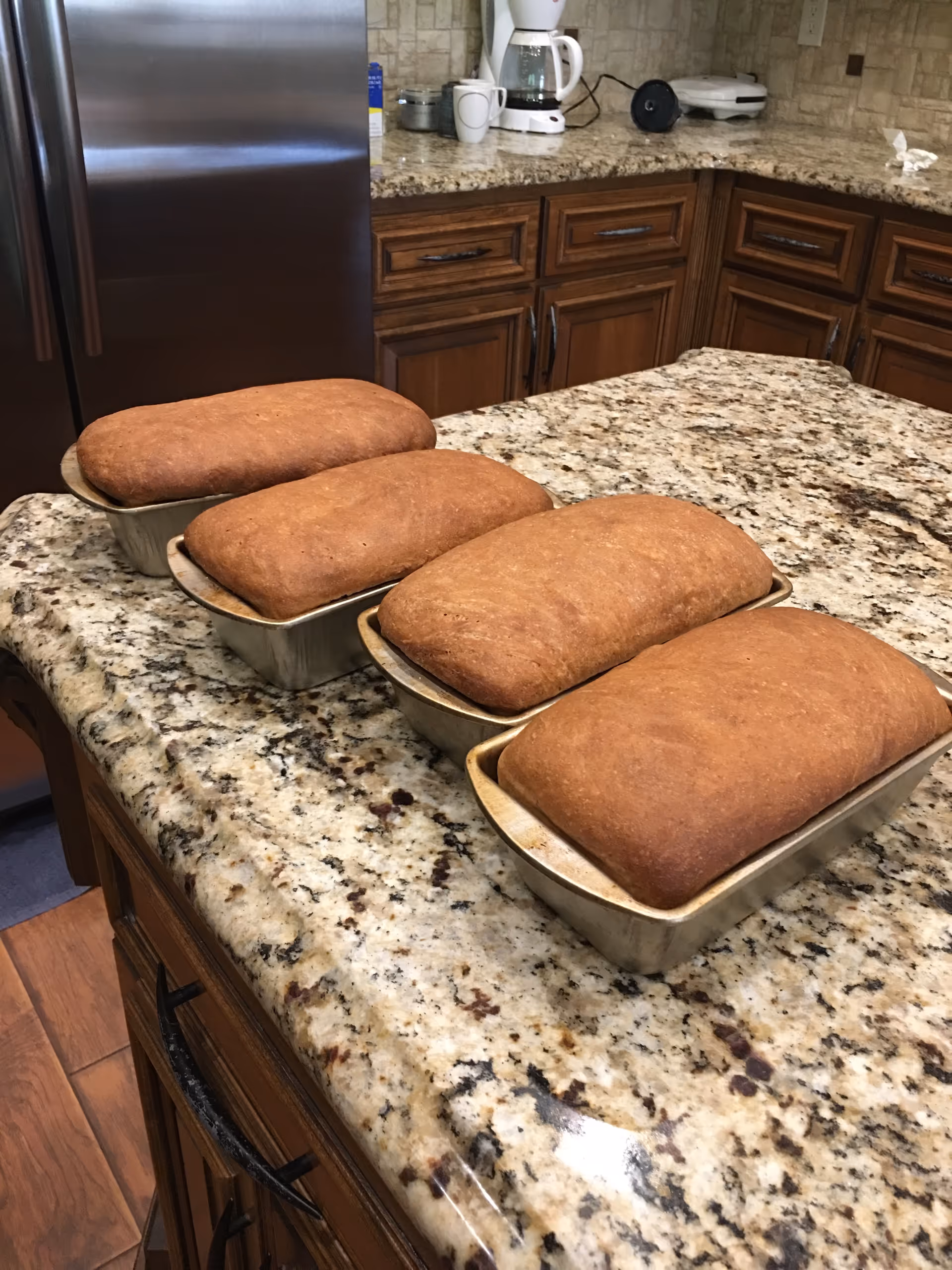 Four freshly baked loaves of bread in metal pans placed on a granite kitchen countertop with wooden cabinets and a stainless steel refrigerator in the background.
