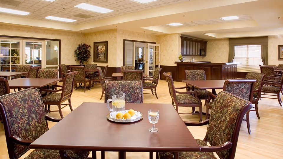 A dining room in a senior living facility with multiple wooden tables and cushioned chairs featuring floral upholstery. On the nearest table, there is a tray with a pitcher of water and lemons, a glass of water, and some lemons. The room has beige walls, a wooden floor, and a serving counter with cabinets in the background. Large windows with blinds allow natural light into the space.