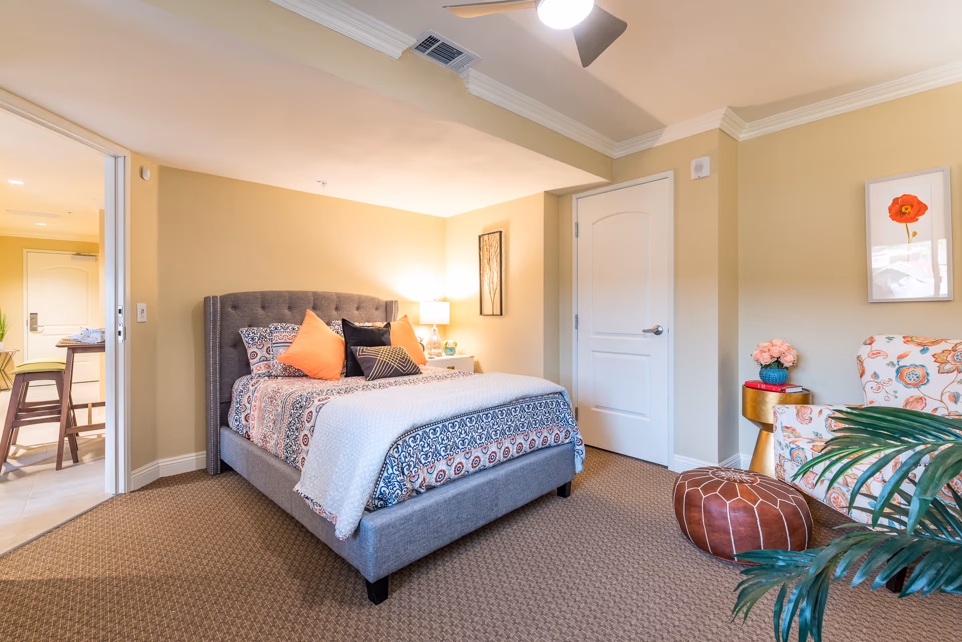 A cozy bedroom in a senior living facility featuring a gray upholstered bed with patterned bedding and orange and black pillows. To the right, there is a floral armchair, a round brown leather ottoman, a small gold side table with a vase of pink flowers, and a framed picture of a red flower on the wall. The room has beige walls, carpeted floor, and a white door. A ceiling fan light is visible above, and an open doorway leads to a kitchen area with bar stools.