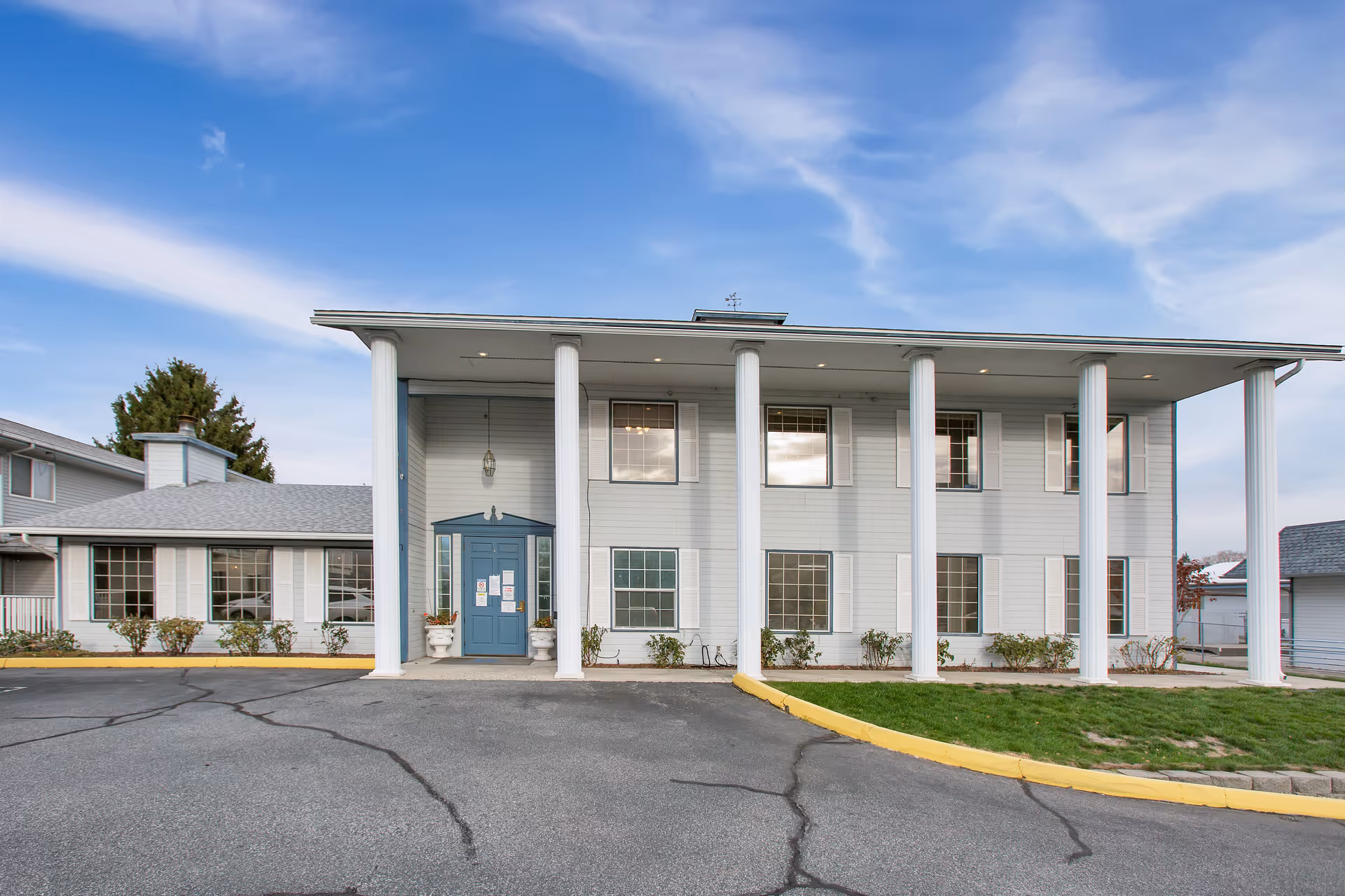 Front exterior view of a two-story senior living facility building with white columns, blue double doors, multiple windows, and a paved driveway with a yellow curb. The sky is partly cloudy.