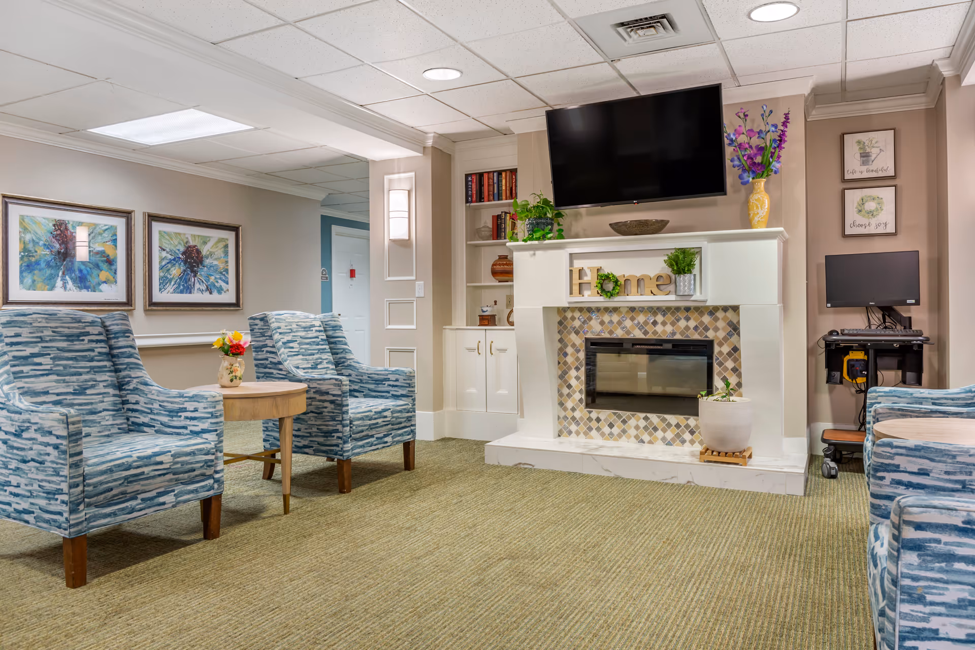Cozy common room with patterned armchairs, a fireplace mantel decorated with plants and a wall-mounted TV.