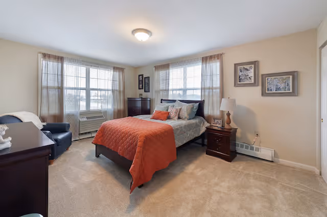 Bright, carpeted bedroom with a bed dressed in an orange quilt, nightstands, a dresser, an armchair, and two large windows.
