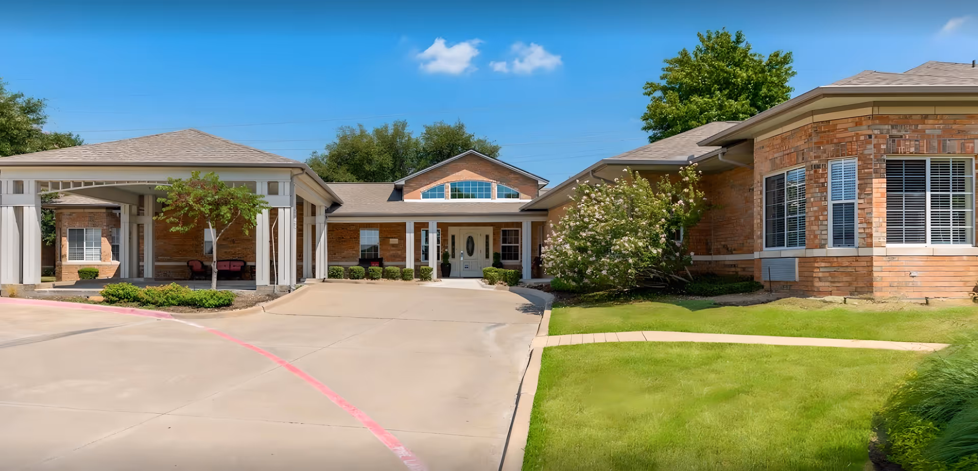 Front entrance of a single-story brick assisted living building with a covered drive-up portico, driveway, and manicured lawn under a blue sky.