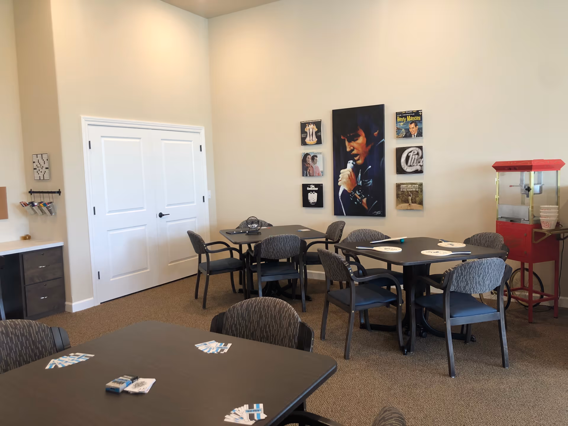 Community dining/activity room with several tables and chairs, wall art on the far wall, and a popcorn machine.