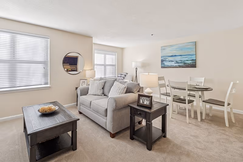 A bright living room and dining area with beige walls and carpet. The living room features a gray loveseat with striped pillows, a dark wooden coffee table with a decorative bowl, and a matching side table with a lamp and framed photo. The dining area has a round wooden table with four white chairs, and a large painting of an ocean scene hangs on the wall. Two windows with white blinds allow natural light into the space.