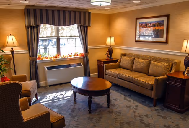 A cozy living room in a senior living facility with a patterned sofa, two armchairs, a round wooden coffee table, two side tables with lamps, a large window with striped curtains, and a framed landscape painting on the wall.
