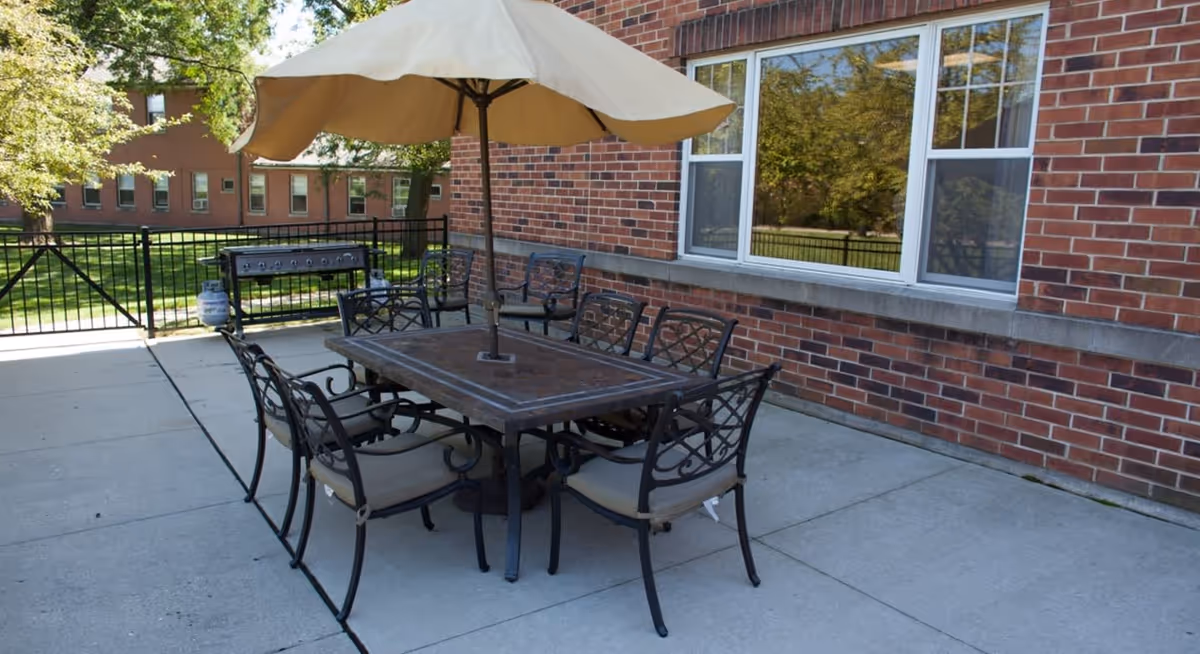 Outdoor patio area with a rectangular metal table and six cushioned chairs arranged around it. A large beige umbrella is positioned in the center of the table. In the background, there is a brick building with windows and a black metal fence enclosing the patio. A grill is also visible near the fence.