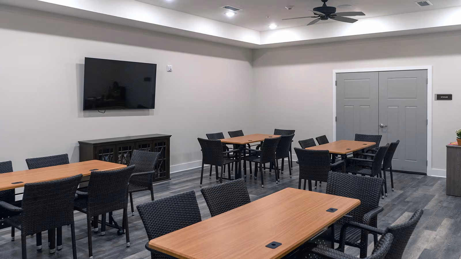 A room with multiple rectangular wooden tables and black wicker chairs arranged around them. A flat-screen TV is mounted on a light gray wall above a dark wooden cabinet. The room has gray double doors labeled 'Storage', a ceiling fan, and recessed lighting.