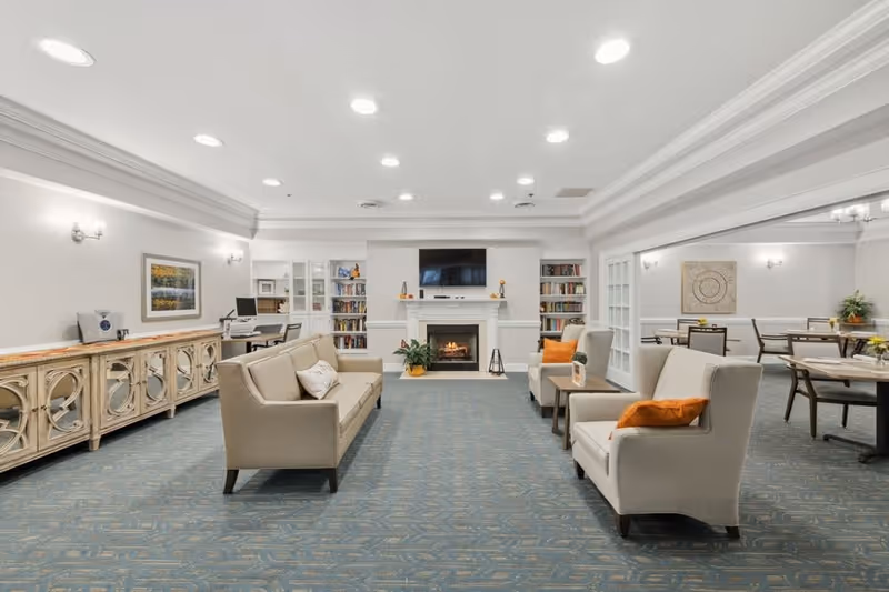 A spacious and well-lit living room area in a senior living facility featuring beige sofas and armchairs with orange cushions, a fireplace with a TV mounted above it, built-in bookshelves on either side, and a sideboard along one wall. Adjacent to the living room is a dining area with tables and chairs, decorated with wall art and plants.