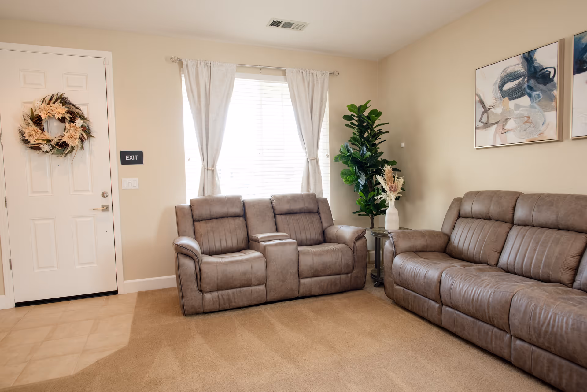 A cozy living room with two brown leather sofas, a window with white curtains, a small round side table with a white vase holding dried flowers, a green potted plant, and a door decorated with a floral wreath. Two abstract paintings hang on the beige wall.