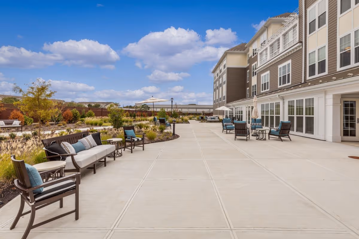 Outdoor patio area at Sunrise of New Dorp featuring multiple seating arrangements with cushioned chairs and benches along a wide concrete walkway. The patio is adjacent to a multi-story building with large windows, under a blue sky with scattered clouds. There are landscaped garden beds with plants and small trees surrounding the seating area.