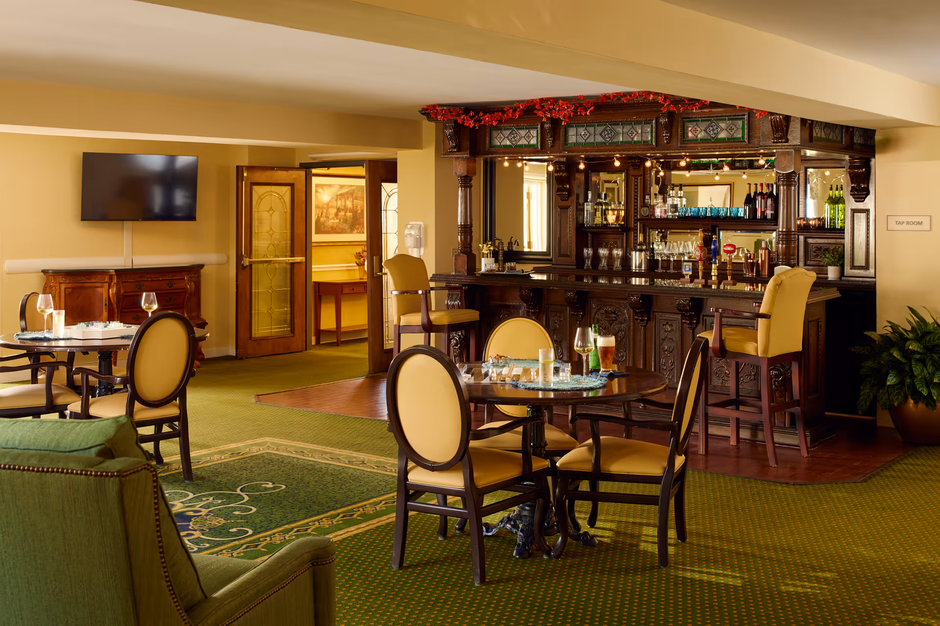A cozy lounge area with a green carpet and several round tables surrounded by yellow cushioned chairs. There is a wooden bar with ornate carvings and a mirror behind it, stocked with glasses and bottles. A green armchair is visible in the foreground, and a flat-screen TV is mounted on the wall. The room has warm lighting and a welcoming atmosphere.