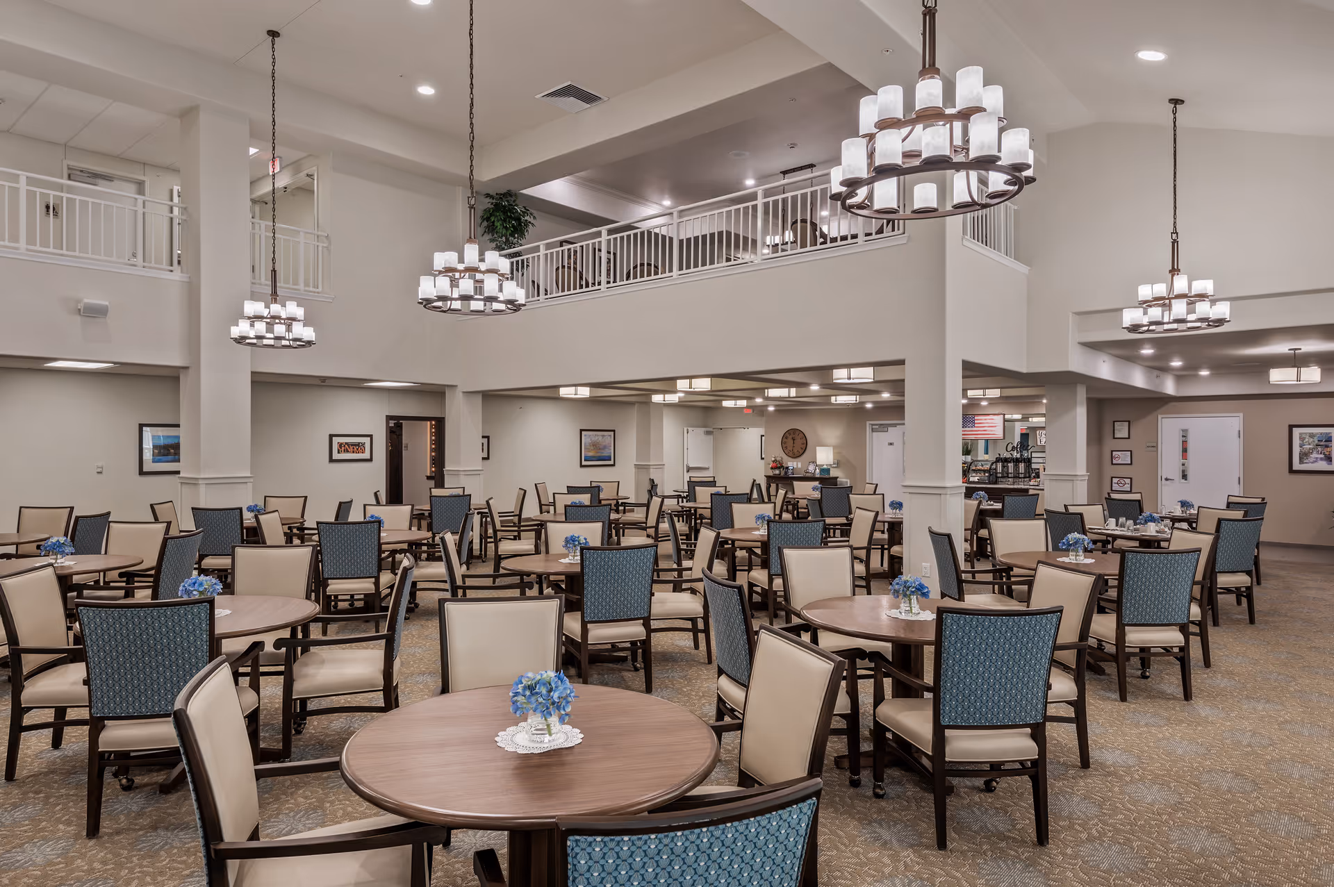 Spacious senior living dining room with multiple round tables and chairs, chandeliers, and an upper balcony.