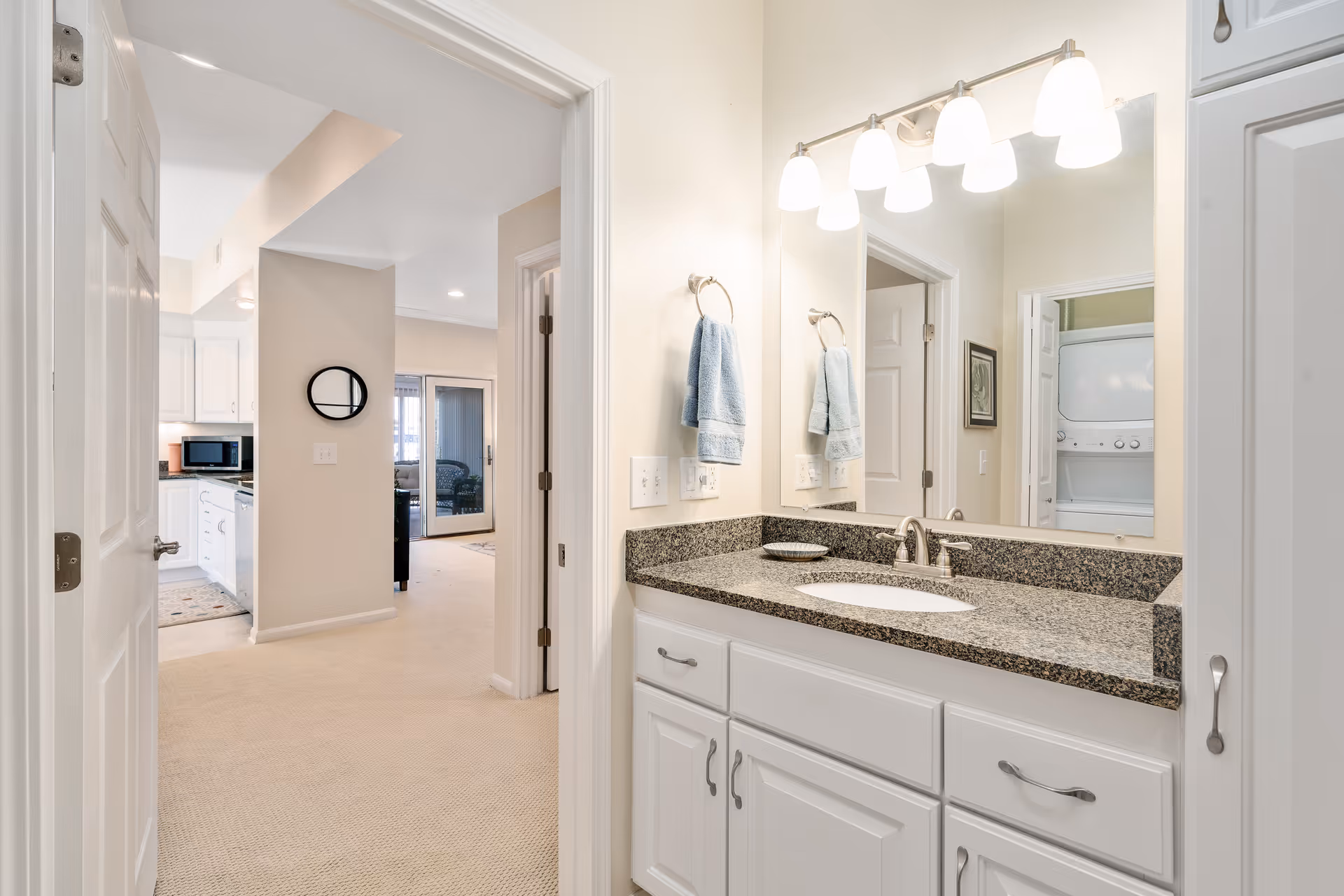 View of a bathroom vanity with a granite countertop, white cabinets, a large mirror, and five light fixtures above. Two blue hand towels hang on towel rings on the wall. The bathroom opens into a hallway leading to a kitchen area with white cabinetry and a microwave, and a living room with sliding glass doors. A stacked washer and dryer unit is visible through a doorway in the hallway.
