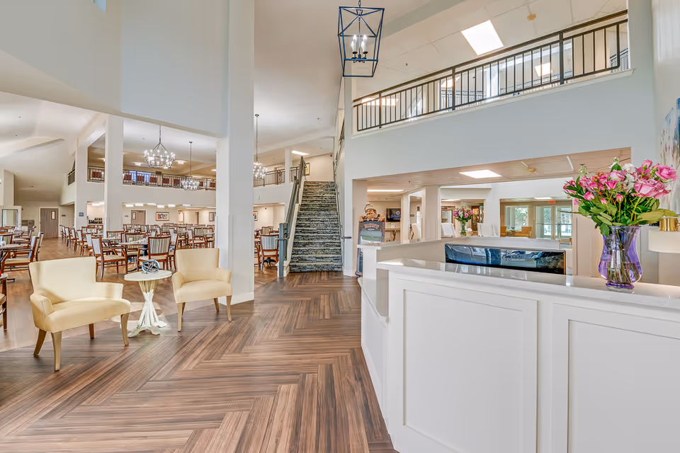 Bright and spacious senior living facility interior with a reception desk on the right adorned with a vase of pink flowers. To the left, there are two beige armchairs with a small round table between them. In the background, there is a large dining area with multiple wooden tables and chairs, chandeliers hanging from the ceiling, and a staircase leading to an upper floor with a railing.