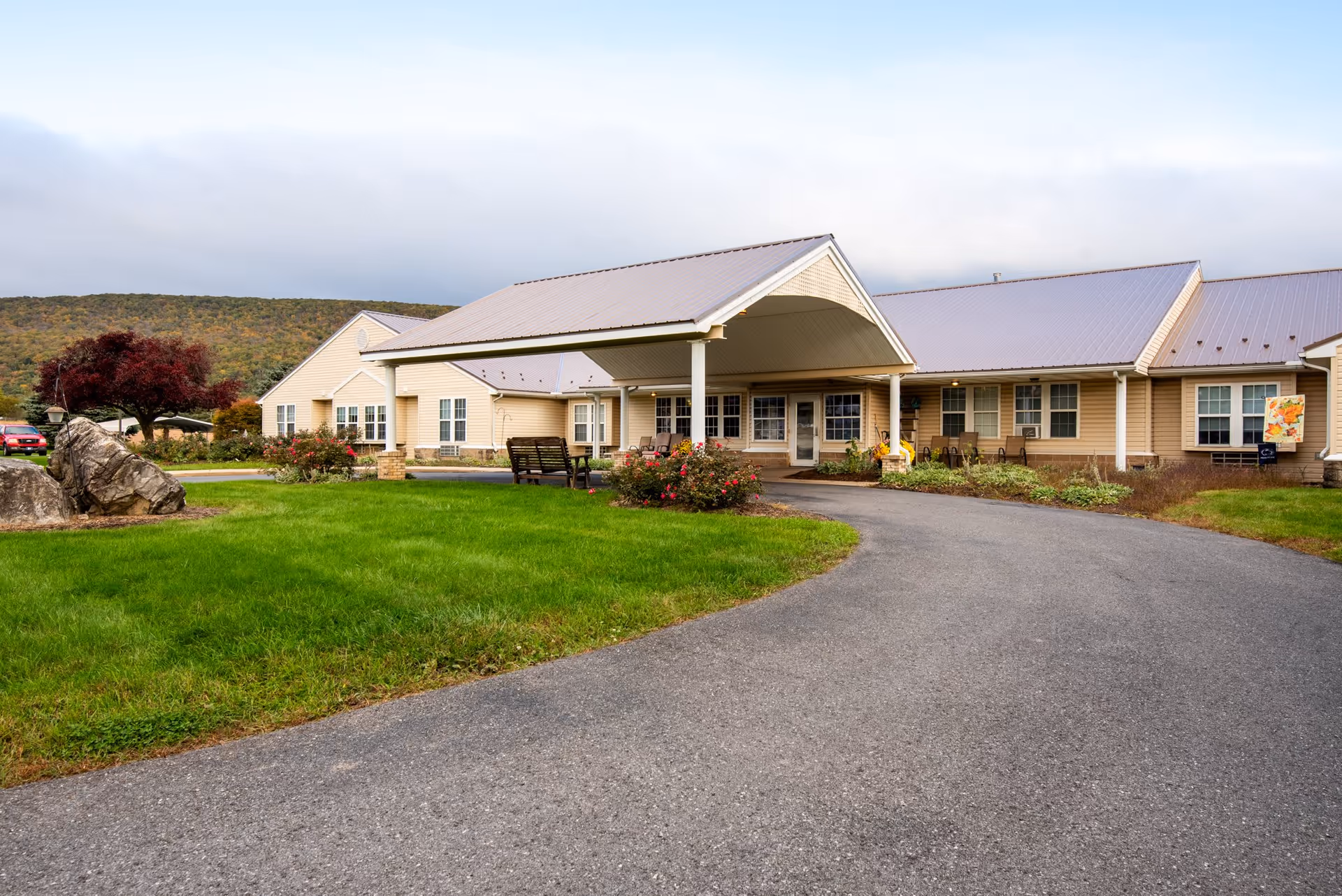 Front exterior of a single-story senior living building with a covered entrance, driveway, lawn, and benches.