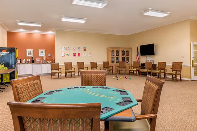 A spacious common room with a green octagonal card table and four wooden chairs in the foreground. In the background, there are multiple wooden chairs arranged along the walls, a cabinet with glass doors, a flat-screen TV mounted on the wall, an American flag, and a refreshment area with a soda machine and countertop. Colorful artwork is displayed on the far wall, and several small colorful balls are scattered on the carpeted floor.