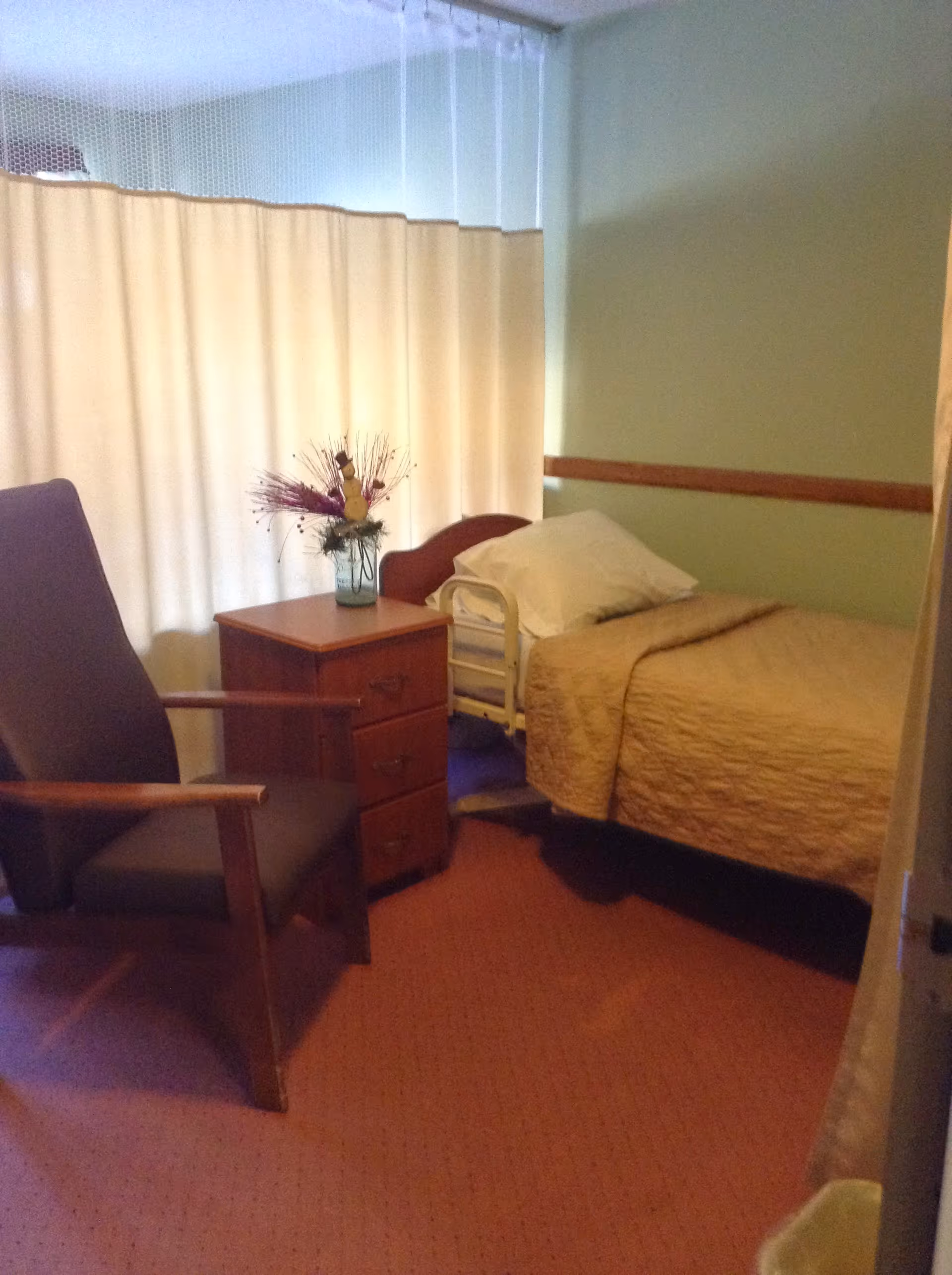 A small senior living bedroom with a single bed covered in a beige quilt, a wooden nightstand with a vase of flowers, and a cushioned armchair. The room has light green walls and a beige curtain covering the window.