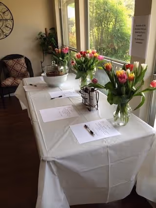 A table covered with a white tablecloth is set up near large windows with a view of greenery outside. On the table are three vases filled with colorful tulips, several sheets of paper with pens placed on top, and a decorative lantern. A chair with a patterned cushion is positioned against the wall to the left, and a small plant is nearby. A sign is posted on the window.