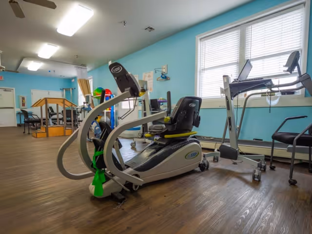 A rehabilitation exercise room with various fitness equipment including a recumbent exercise bike, a treadmill, and parallel bars. The room has light blue walls, large windows with blinds, and wooden flooring.