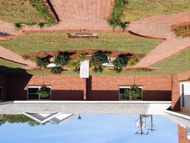 Outdoor garden area with brick pathways, green grass, and flower beds in front of a brick building with windows and white trim under a clear blue sky.
