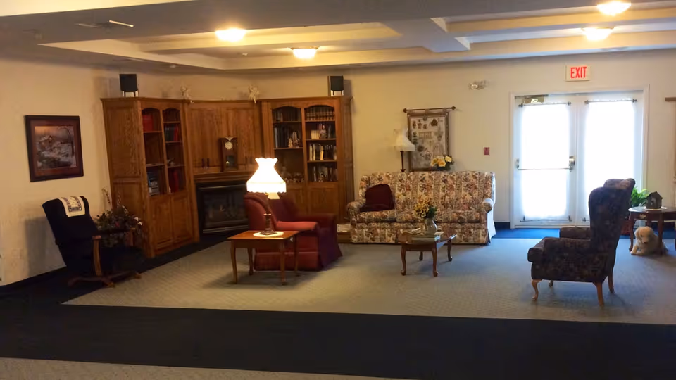 A cozy living room area with a floral-patterned sofa, two armchairs, a wooden coffee table, and a side table with a lamp. There is a wooden cabinet with books and decorative items, a fireplace, and a framed picture on the wall. Double glass doors with an exit sign above are visible in the background.