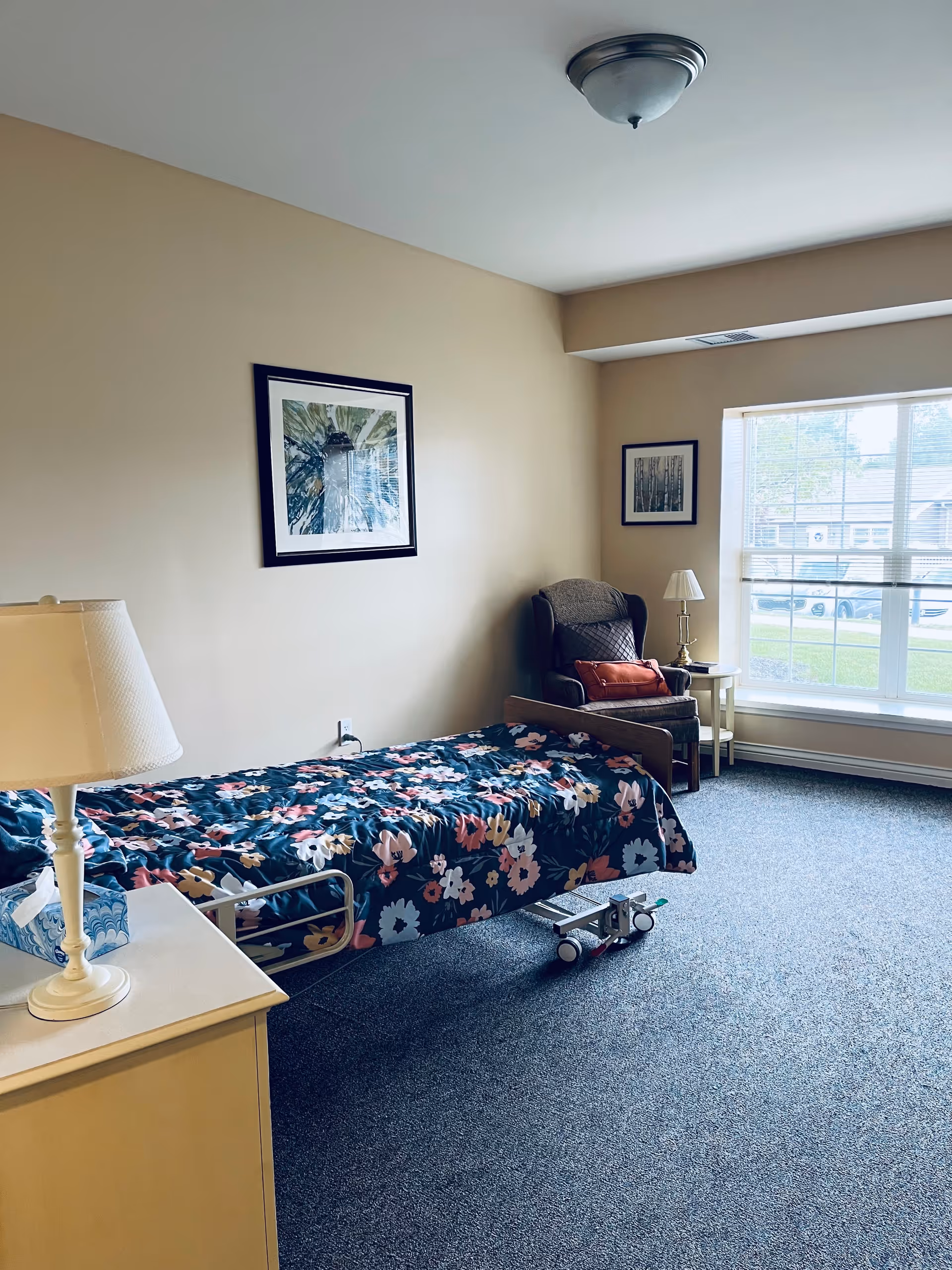 A simple bedroom in a senior living facility with a single bed covered in a floral blanket, a bedside table with a lamp and tissue box, a comfortable armchair with a pillow, a small side table with a lamp, and a large window letting in natural light. Two framed pictures hang on the beige walls.