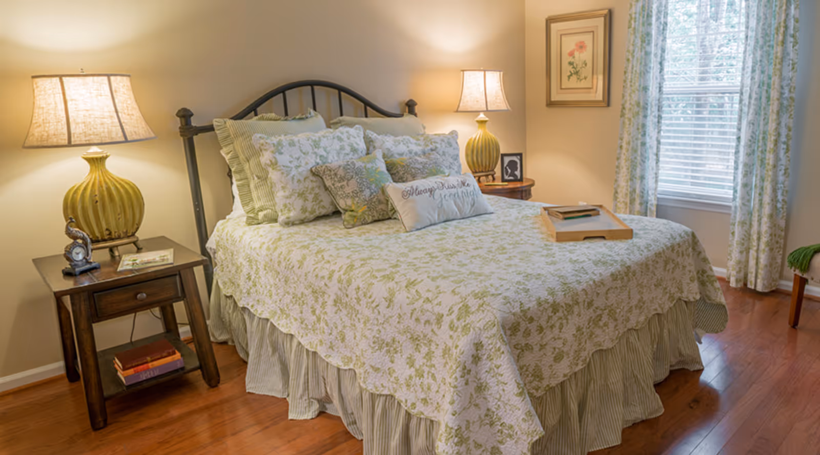 A cozy bedroom with a bed covered in a green and white floral bedspread and multiple pillows. There are two bedside tables with matching lamps, a framed floral picture on the wall, a window with floral curtains, and a wooden floor.