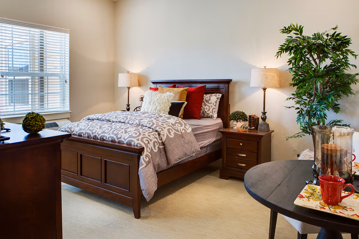 A cozy bedroom with a wooden bed frame, patterned bedding, and multiple decorative pillows. There are two bedside tables with lamps and small plants, a window with blinds, a wooden dresser, a round table with a red mug and decorative items, and a large green potted plant in the corner.