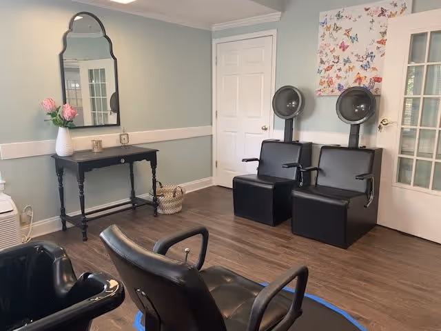Interior of a salon area in a senior living facility with two black hair dryer chairs against a wall, a black styling chair in the foreground, a black table with a mirror above it, a vase with pink flowers, and a butterfly-themed artwork on the wall.