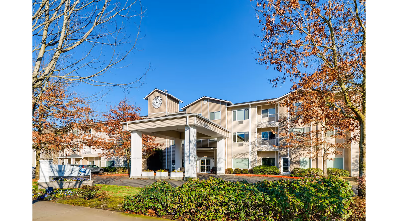 Exterior view of a three-story senior living facility building with beige siding and a clock tower above the entrance. The entrance has a covered drop-off area supported by white columns. Trees with autumn-colored leaves and green shrubs surround the building under a clear blue sky.