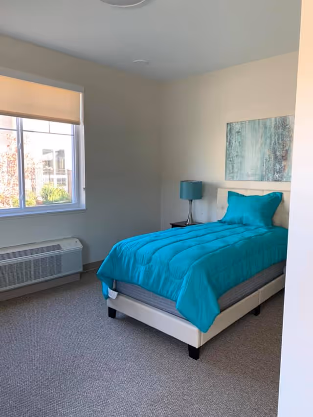 A small bedroom with a single bed featuring a bright turquoise comforter and matching pillow. Next to the bed is a nightstand with a turquoise lamp. A large window with a beige roller shade lets in natural light, and a piece of abstract art hangs on the wall above the bed. The room has beige walls and carpeted flooring.