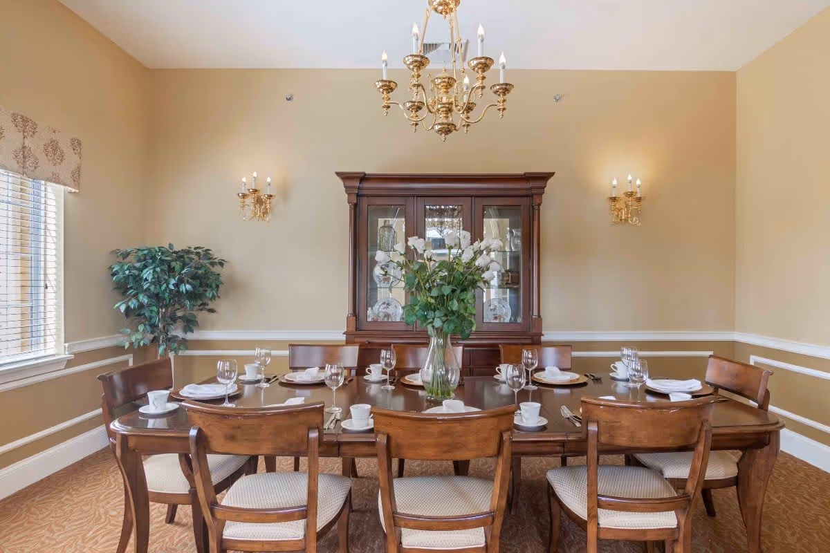 A formal dining room with a wooden dining table set for eight people. The table has white plates, cups, wine glasses, and silverware arranged neatly. A large vase with white flowers is placed in the center of the table. Behind the table is a wooden china cabinet displaying decorative plates and glassware. The room has beige walls with white trim, a window with blinds and a patterned valance, a potted plant in the corner, two wall sconces with candles, and a gold chandelier hanging from the ceiling.