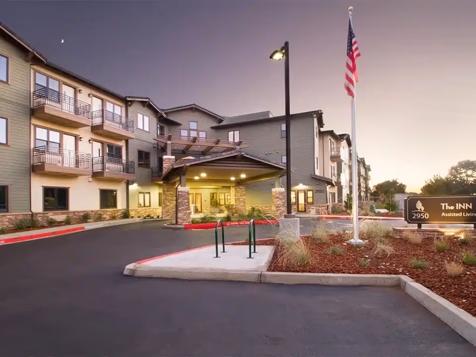 Front entrance of a multi-story assisted living building with a lit porte-cochere, American flag, and landscaping at dusk.